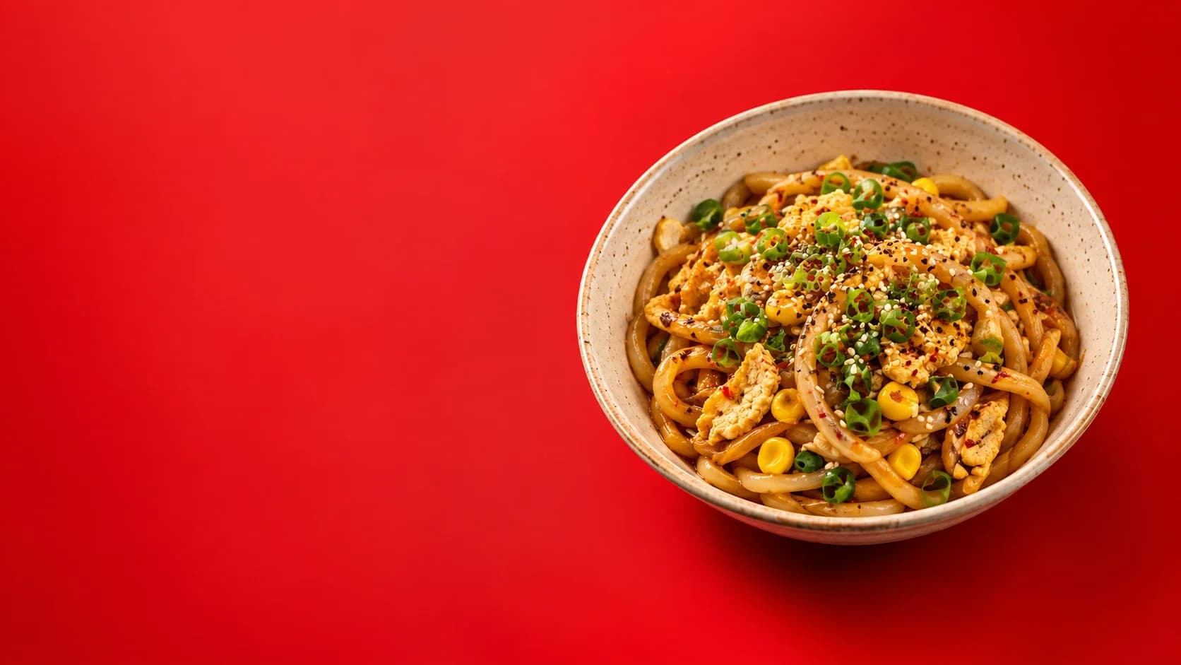 Bowl of chilly garlic udon with tofu, corn, spring onion and sesame on a red background