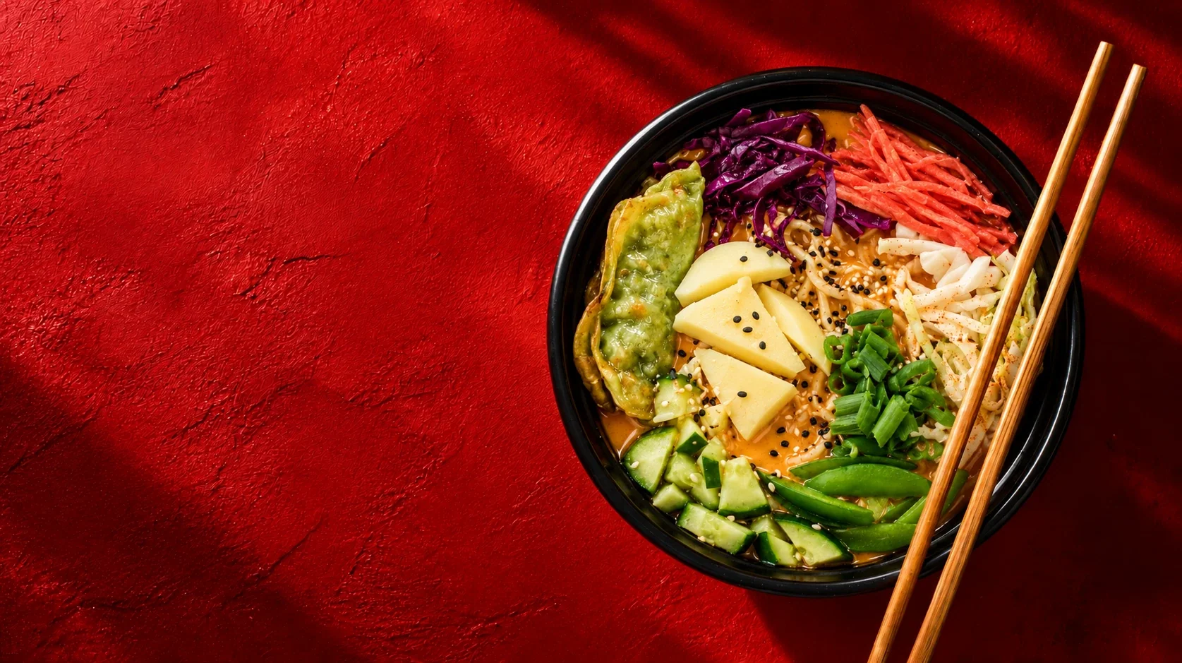 Bowl of creamy SATO ramen with cheese, a dumpling and fresh vegetables, chopsticks resting on top, on a red table